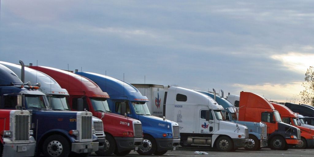 A line of 18 wheel big rig semi trucks parked side by side in a over night truck stop fashion no obvious logos or color scemes very genreric against a sun set sky